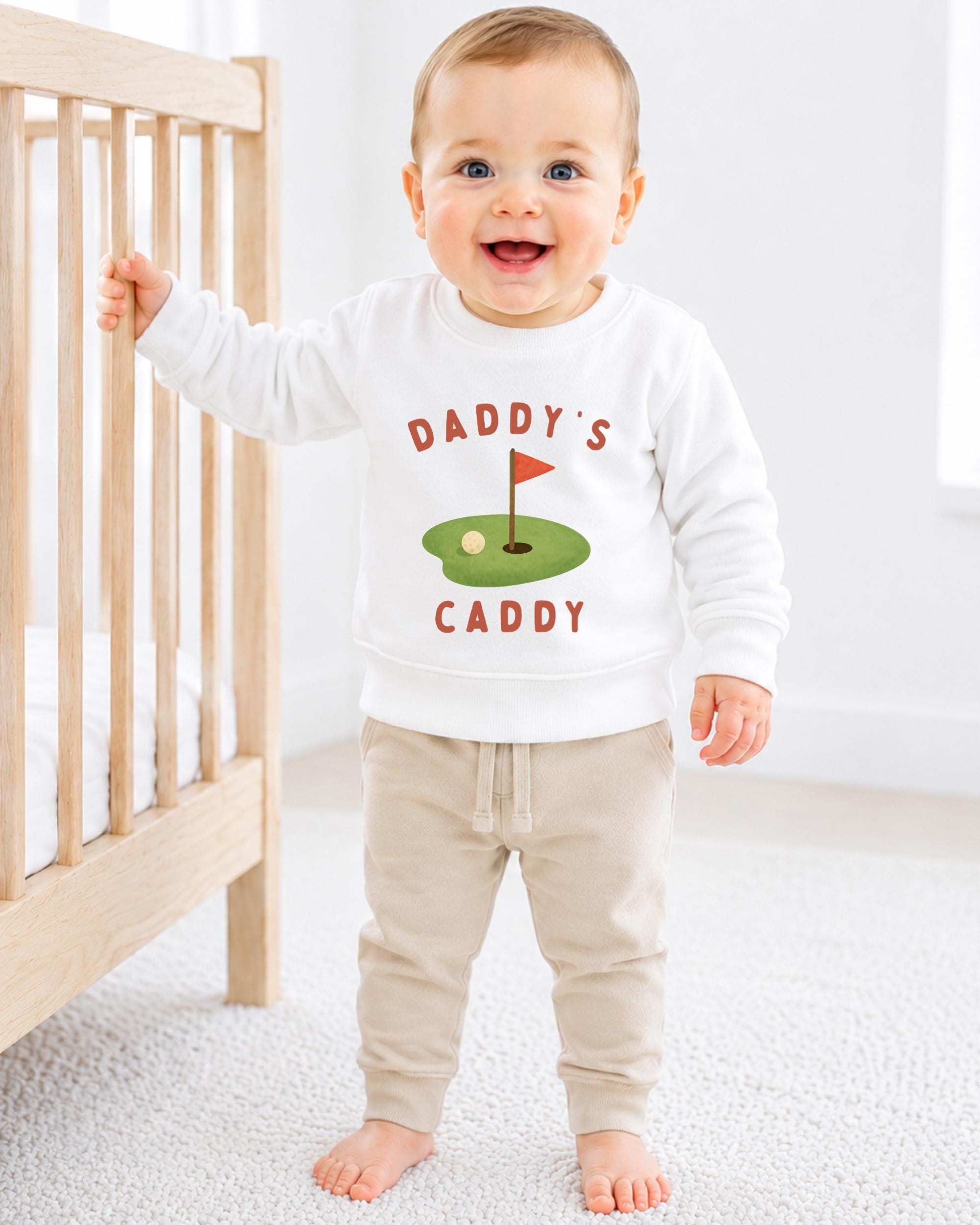 A smiling baby boy wearing a white sweatshirt with a golf hole and flag design, standing in front of a wooden crib.