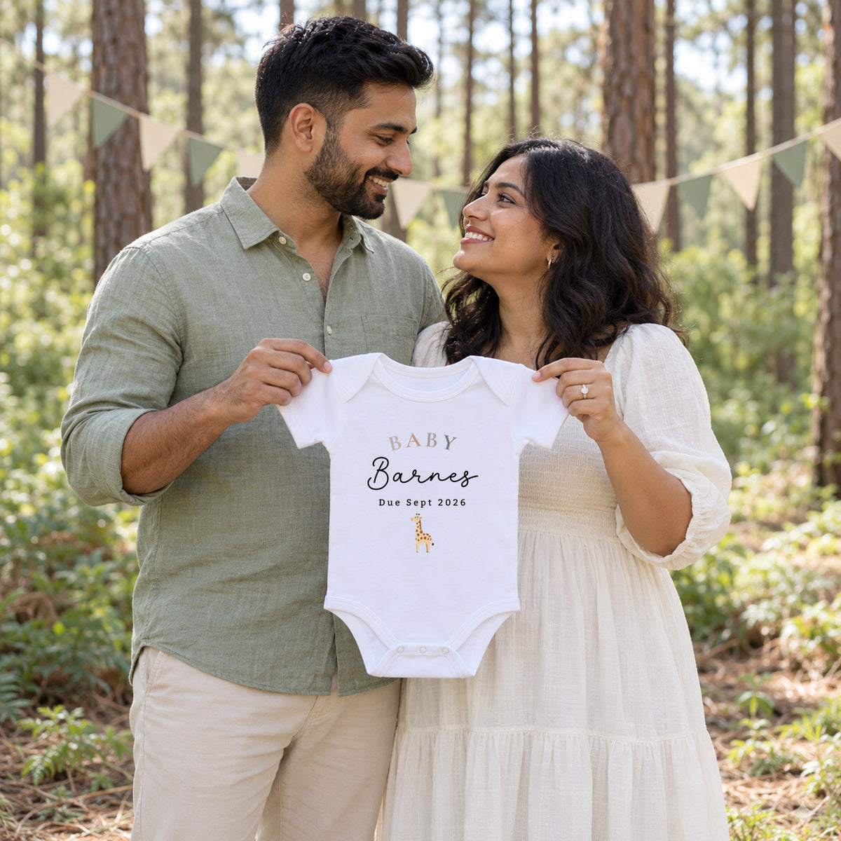 Couple smiling at each other in a pine woodland while holding a white BABY Barnes Due Sept 2026 Baby Vest with a giraffe, with cream and sage bunting behind.