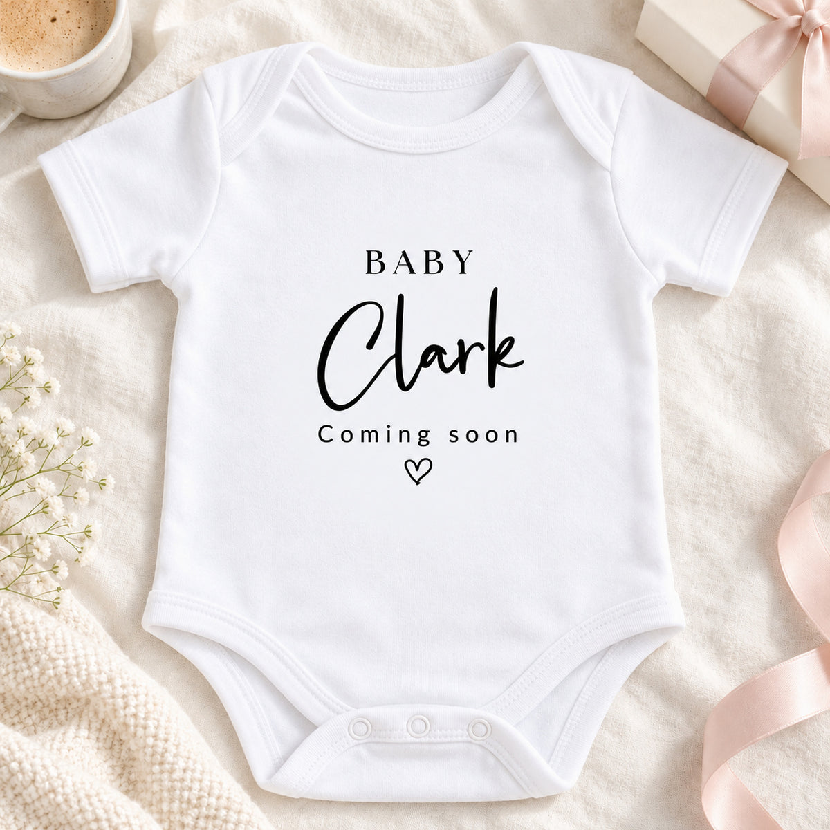 Square pregnancy announcement image of a white Baby Clark Coming Soon Baby Vest with script surname and heart, styled on beige linen with a coffee cup, gift box and ribbon.