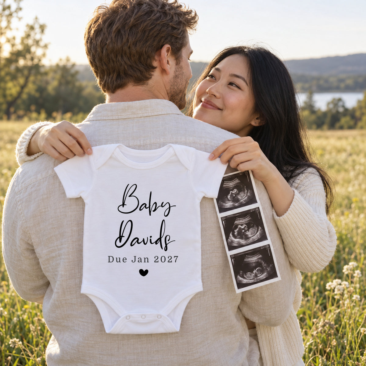 Couple embracing in a meadow, with the woman holding a white Baby Davids Due Jan 2027 Baby Vest with a filled heart against her partner's back, beside a strip of scan photos.