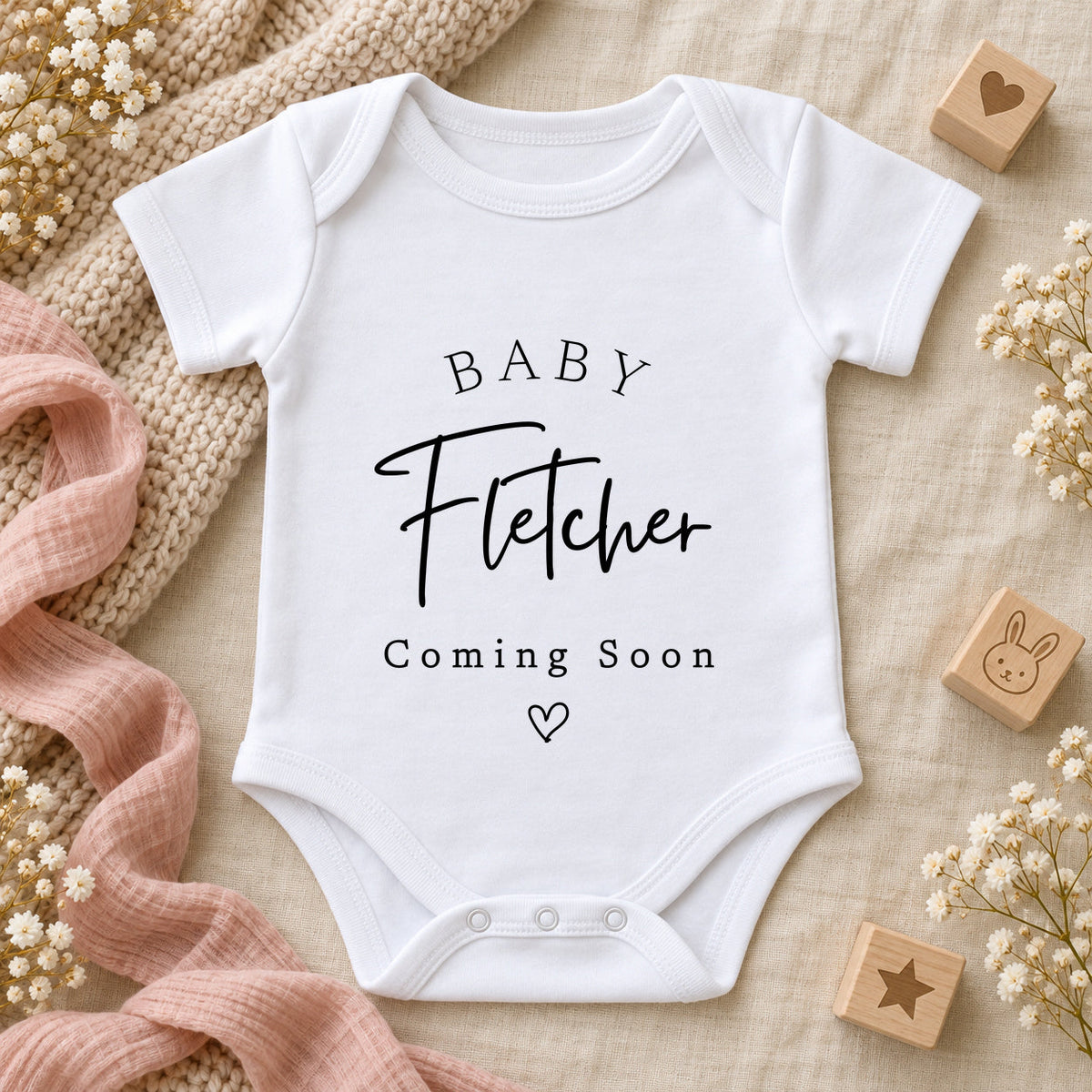 White Baby Vest printed with BABY Fletcher Coming Soon and an outline heart, styled with ivory knitwear, blush muslin, wooden baby blocks and delicate white flowers.
