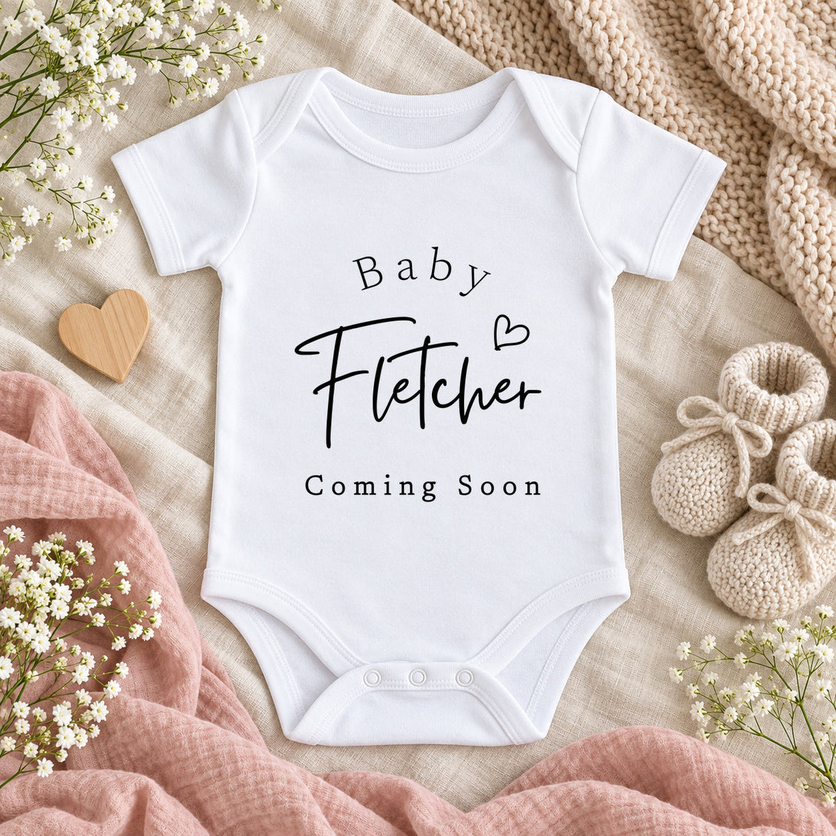 White Baby Vest printed with Baby Fletcher, a heart and Coming Soon, styled on ivory linen with beige knitwear, blush muslin, white flowers, a wooden heart and knitted booties.