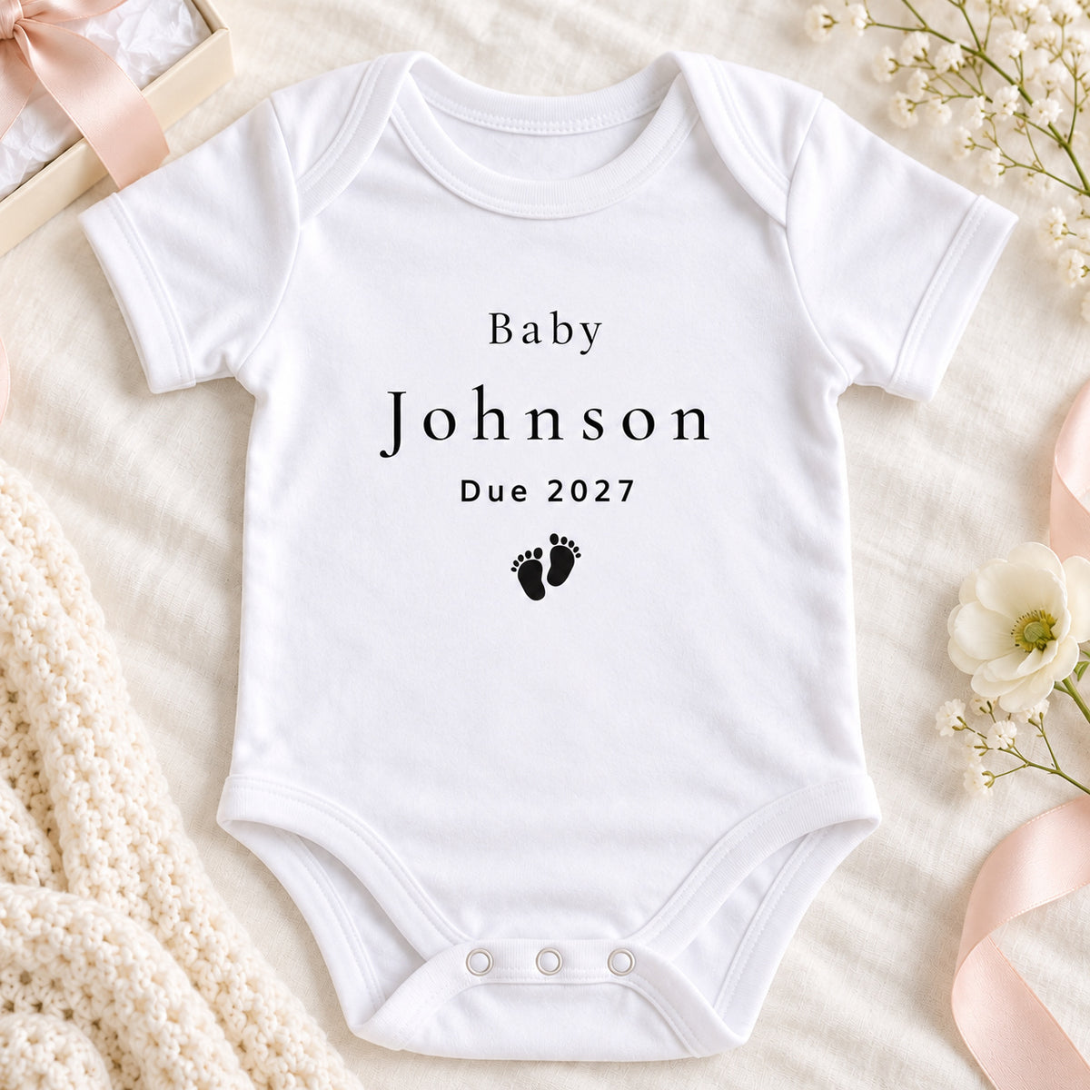 Square pregnancy announcement image of a white Baby Johnson Due 2027 Vest with footprint print, styled beside a scan photo and kraft gift box on beige linen.