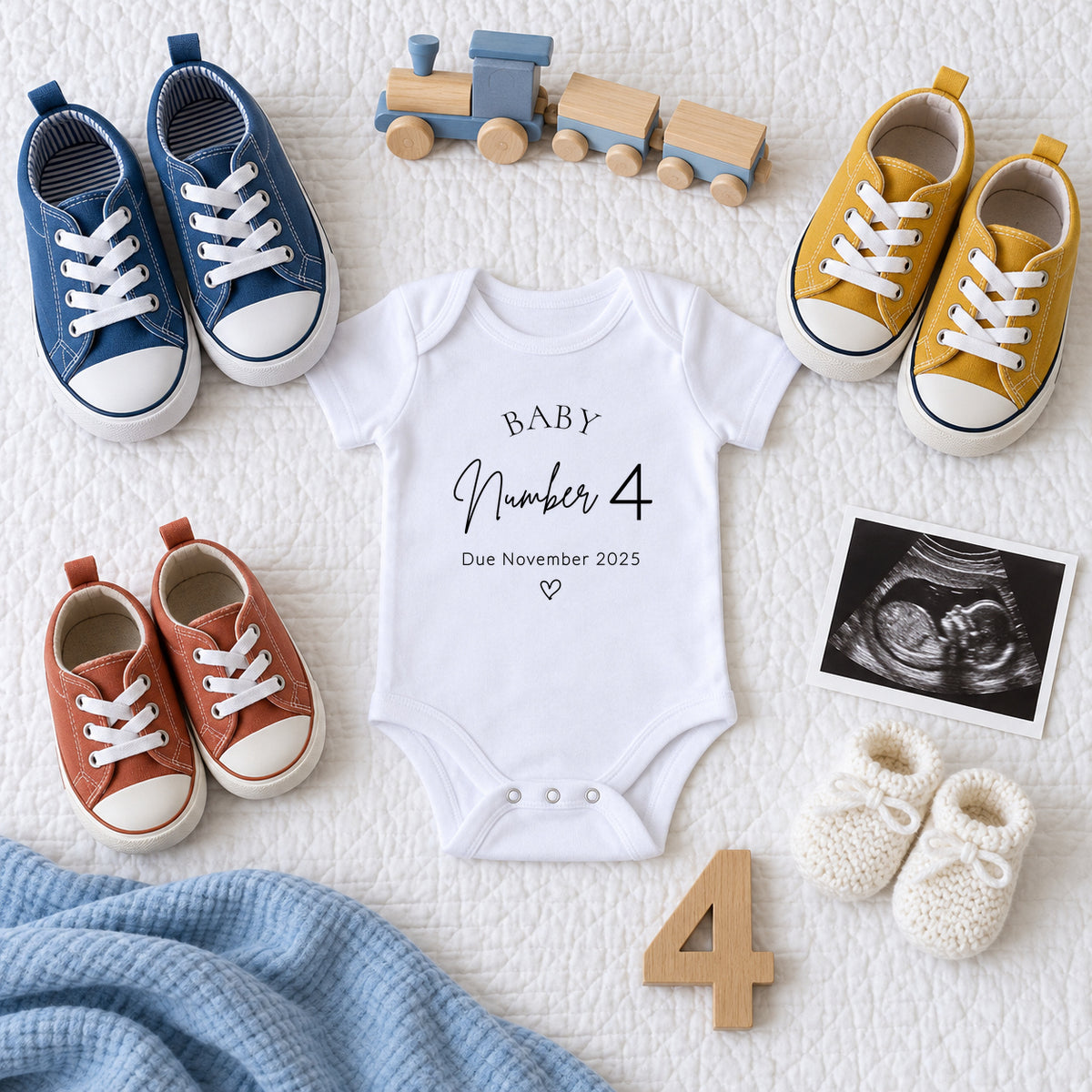 White Baby Number 4 vest reading Due November 2025, surrounded by three pairs of blue, mustard and terracotta children's shoes, newborn booties, a scan photo, wooden train and number four.