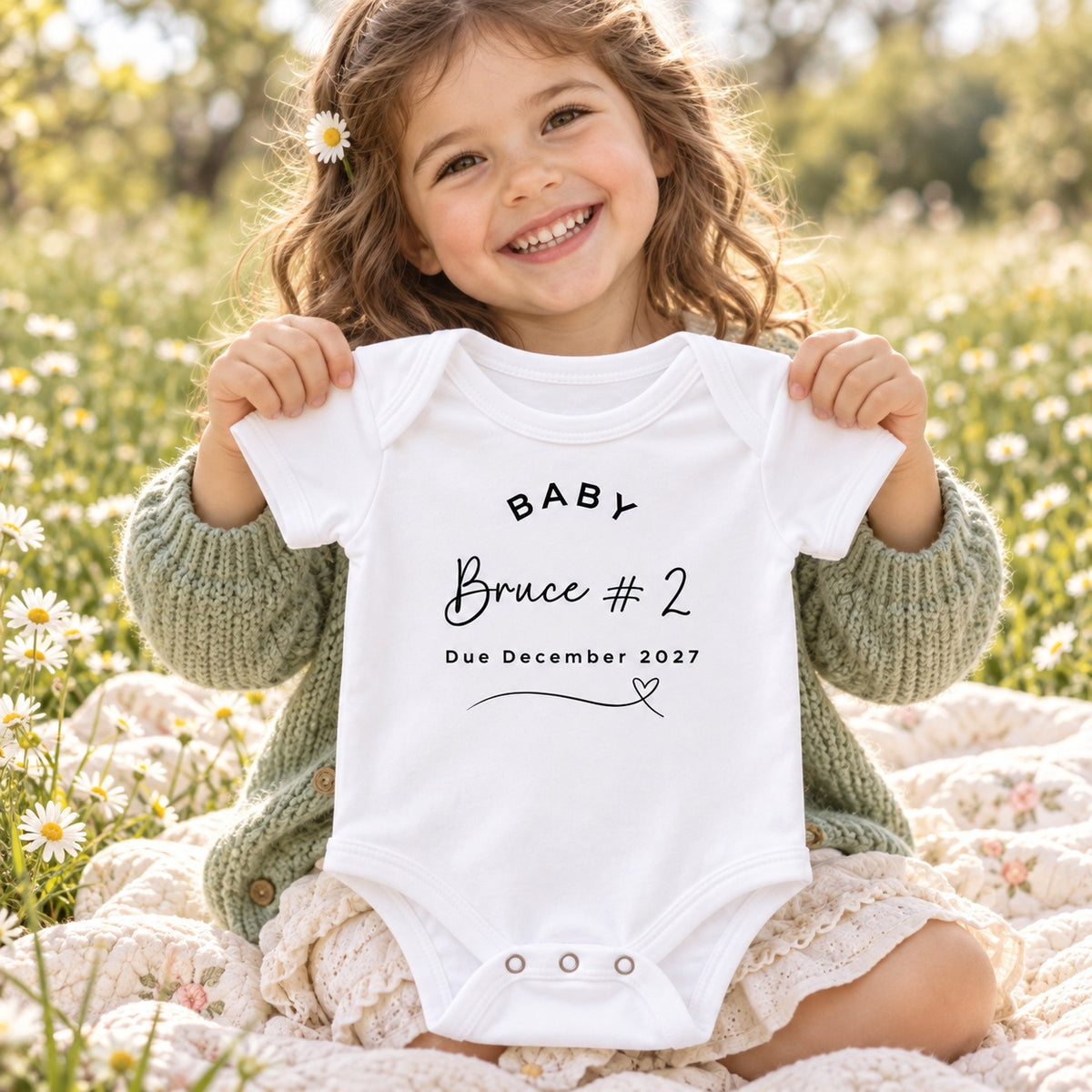 Smiling couple holding a white Baby Vest towards the camera, printed with BABY Bruce #2, Due December 2027 and a heart flourish, in a bright neutral living room.