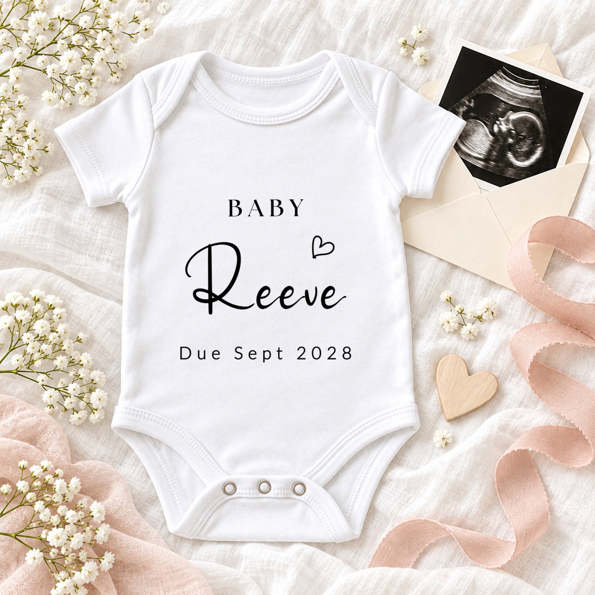 White Baby Vest printed with BABY Reeve, a small heart and Due Sept 2028, styled with a scan photo, cream envelope, blush ribbon, wooden heart and white flowers.