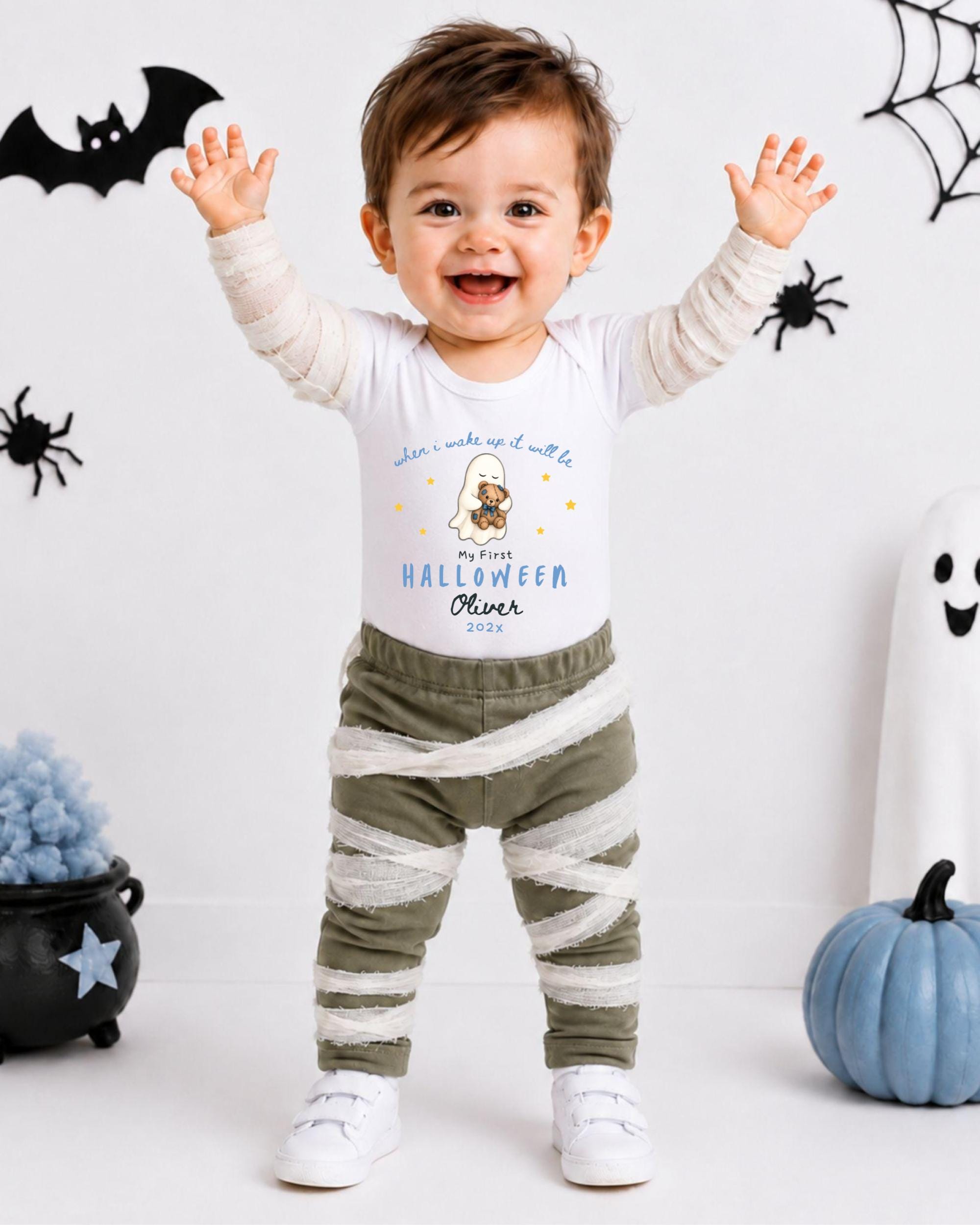 A smiling baby boy wearing a Halloween-themed outfit with a ghost and spider decoration, standing in front of Halloween decorations.
