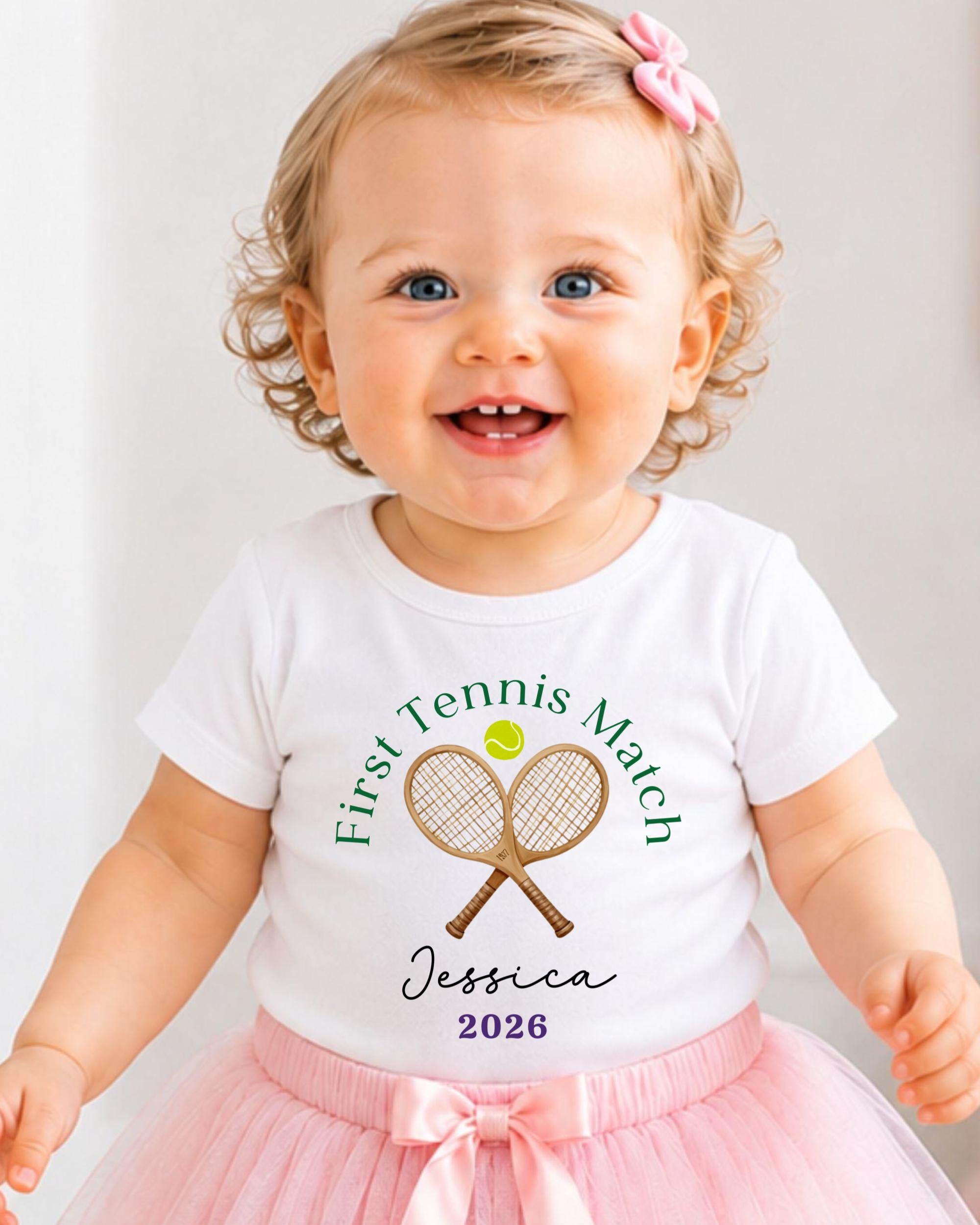 A smiling baby girl wearing a white t-shirt with a tennis racket and ball design, and a pink bow in her hair.