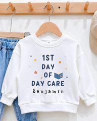 A white sweatshirt with the text "1st DAY OF DAY CARE" printed on it, along with a small illustration of a book and stars.