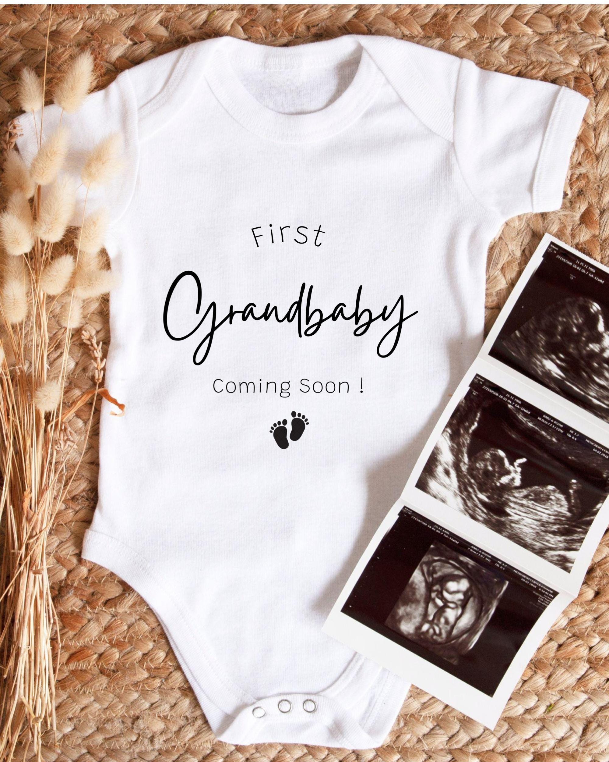 A white onesie with the text "First Grandbaby Coming Soon!" printed on it, along with a pair of footprints. The onesie is placed next to a set of three black and white images, which appear to be ultrasound images of a baby.