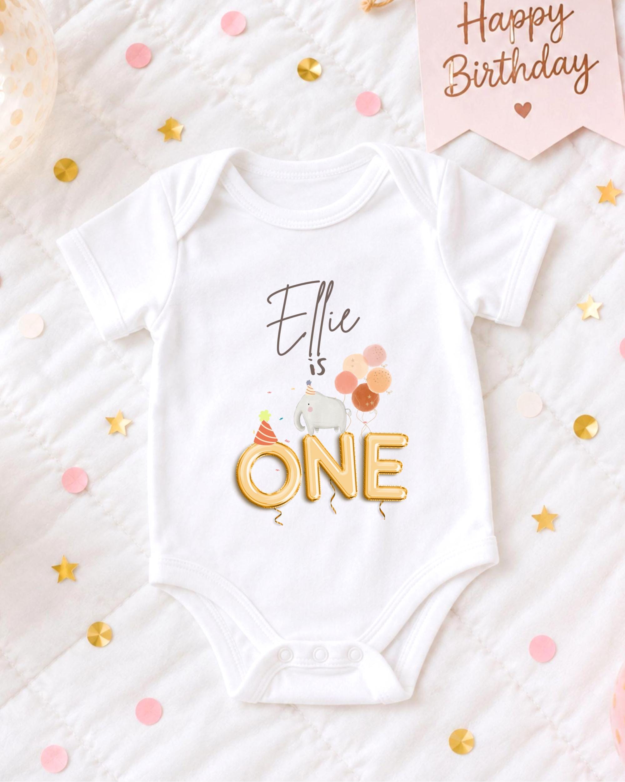 A white onesie with the text "Ellie is one" printed on it, along with a small elephant and balloons, is displayed on a bed surrounded by confetti.