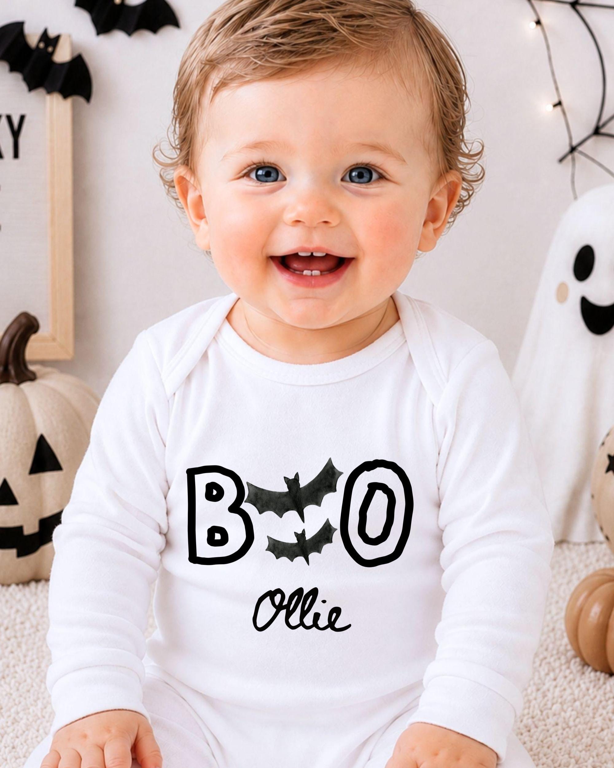 A smiling baby boy wearing a white onesie with the text "BOO OLLIE" printed on it, sitting in front of Halloween decorations.
