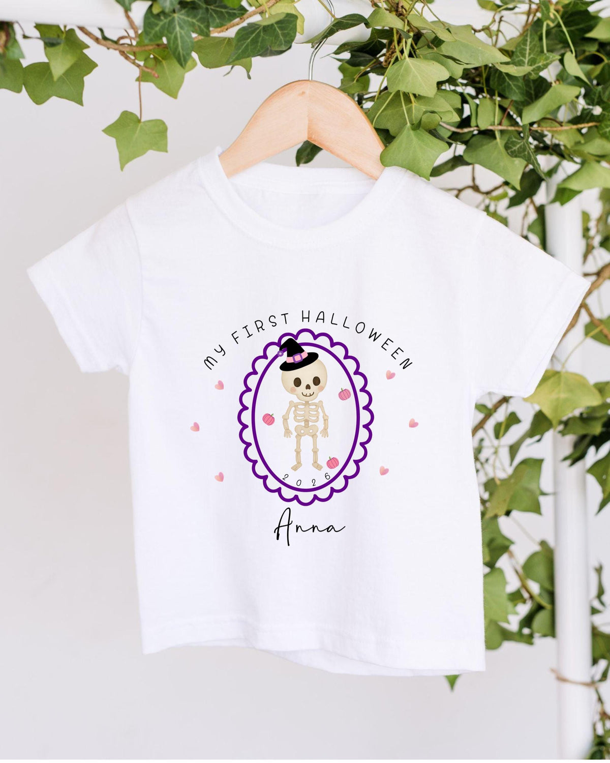 A white t-shirt with a Halloween-themed graphic design hanging on a wooden hanger. The design features a skeleton wearing a witch's hat and surrounded by hearts, with the text "My first Halloween Anna" displayed above the image.