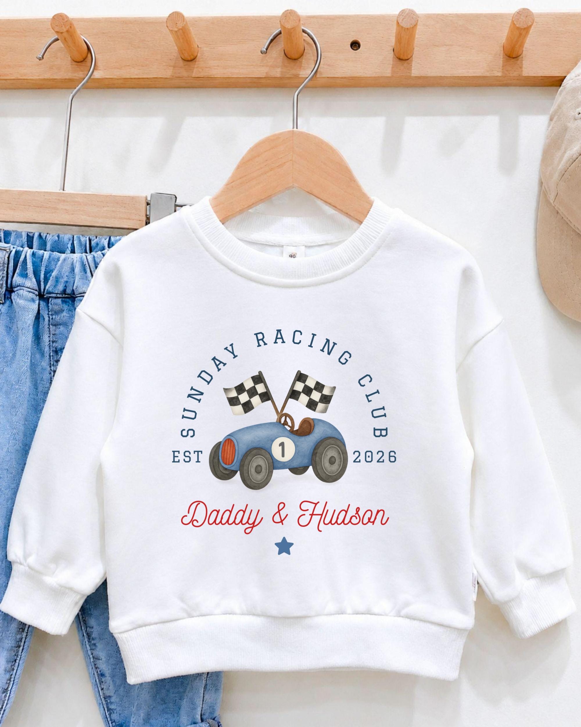 A white sweatshirt with a graphic of a race car and the text "Sunday Racing Club Est. Daddy & Hudson" printed on it, hanging on a wooden hanger.