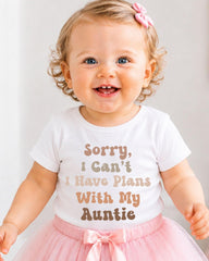A smiling baby girl wearing a white t-shirt with text and a pink bow in her hair.
