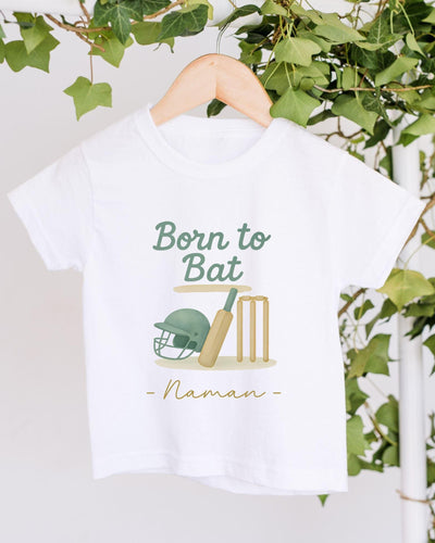 A white t-shirt with a graphic design featuring a cricket helmet, bat, and wickets, along with the text "Born to Bat" and "Naman" printed on it.