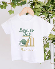 A white t-shirt with a graphic design featuring a cricket helmet, bat, and wickets, along with the text "Born to Bat" and "Naman" printed on it.