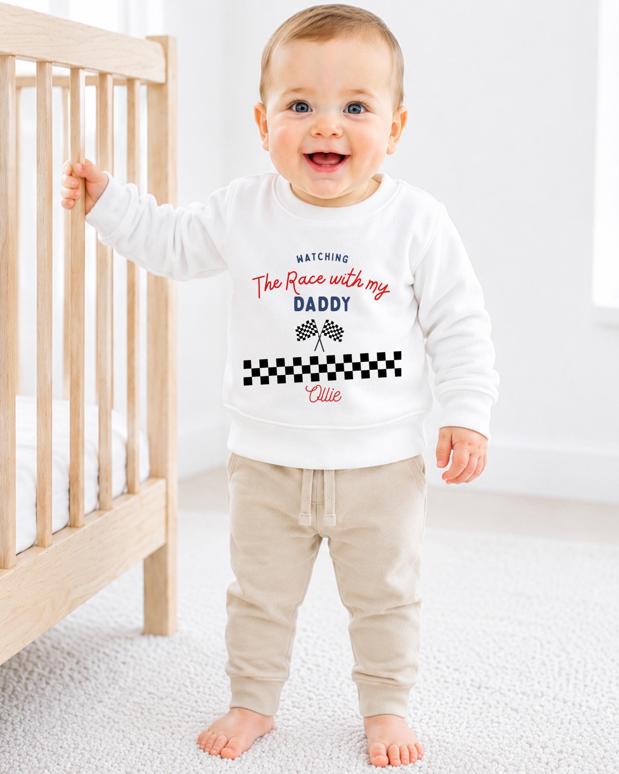 A smiling baby boy wearing a white sweatshirt with a racing theme and text, standing next to a wooden crib.