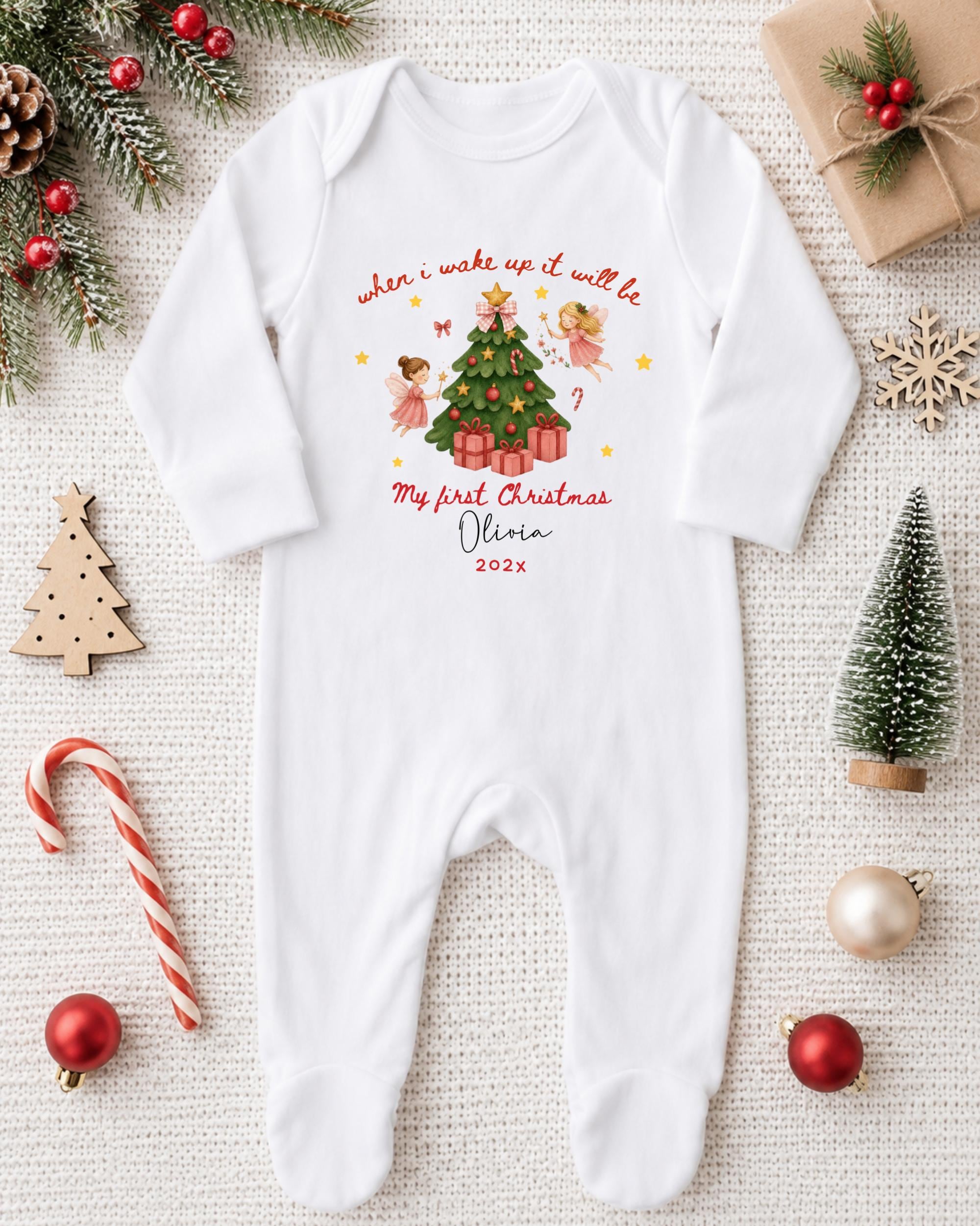 A white onesie with a Christmas-themed design, including a Christmas tree, angels, and presents, is displayed on a white surface alongside various Christmas decorations and gifts.
