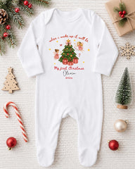 A white onesie with a Christmas-themed design, including a Christmas tree, angels, and presents, is displayed on a white surface alongside various Christmas decorations and gifts.