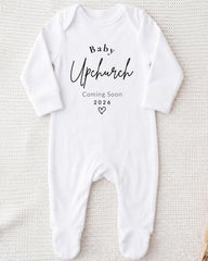 A white onesie with the text "Baby Upchurch" printed on it, along with the text "Coming Soon 2026" below it.