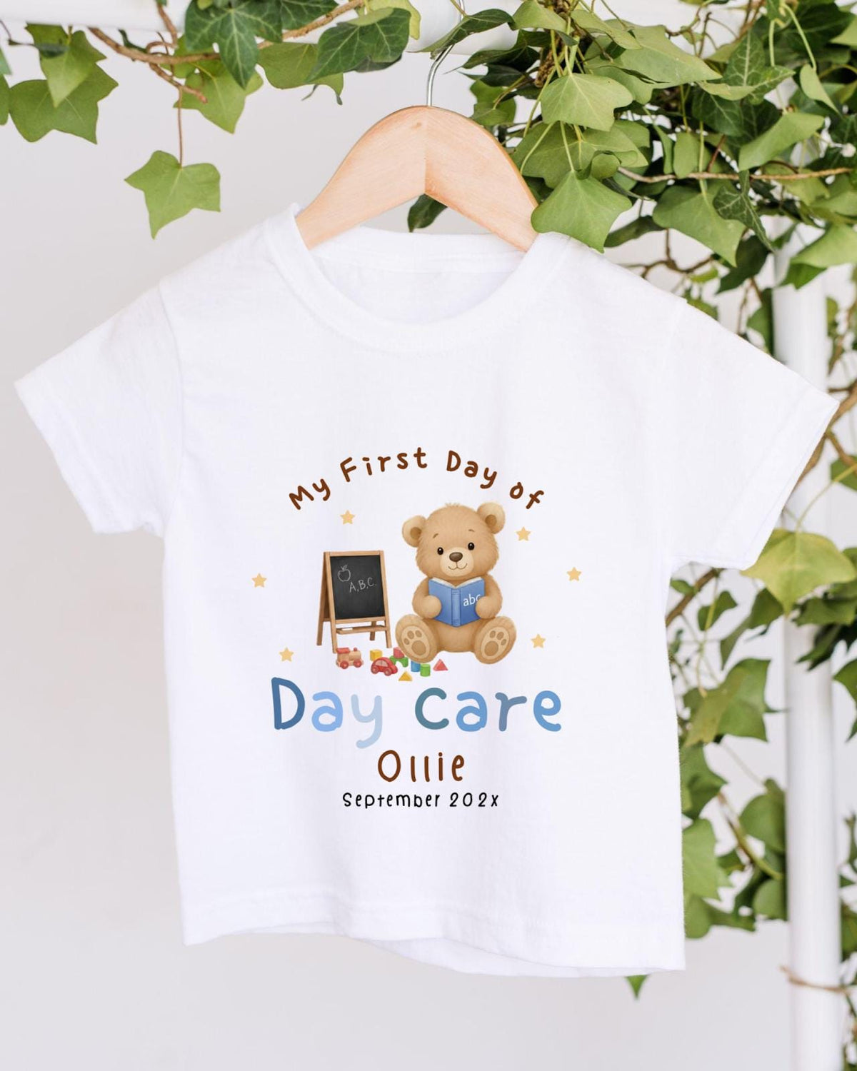 A white t-shirt with a teddy bear and a chalkboard, along with the text "My First Day of Daycare Ollie September 2023" printed on it.