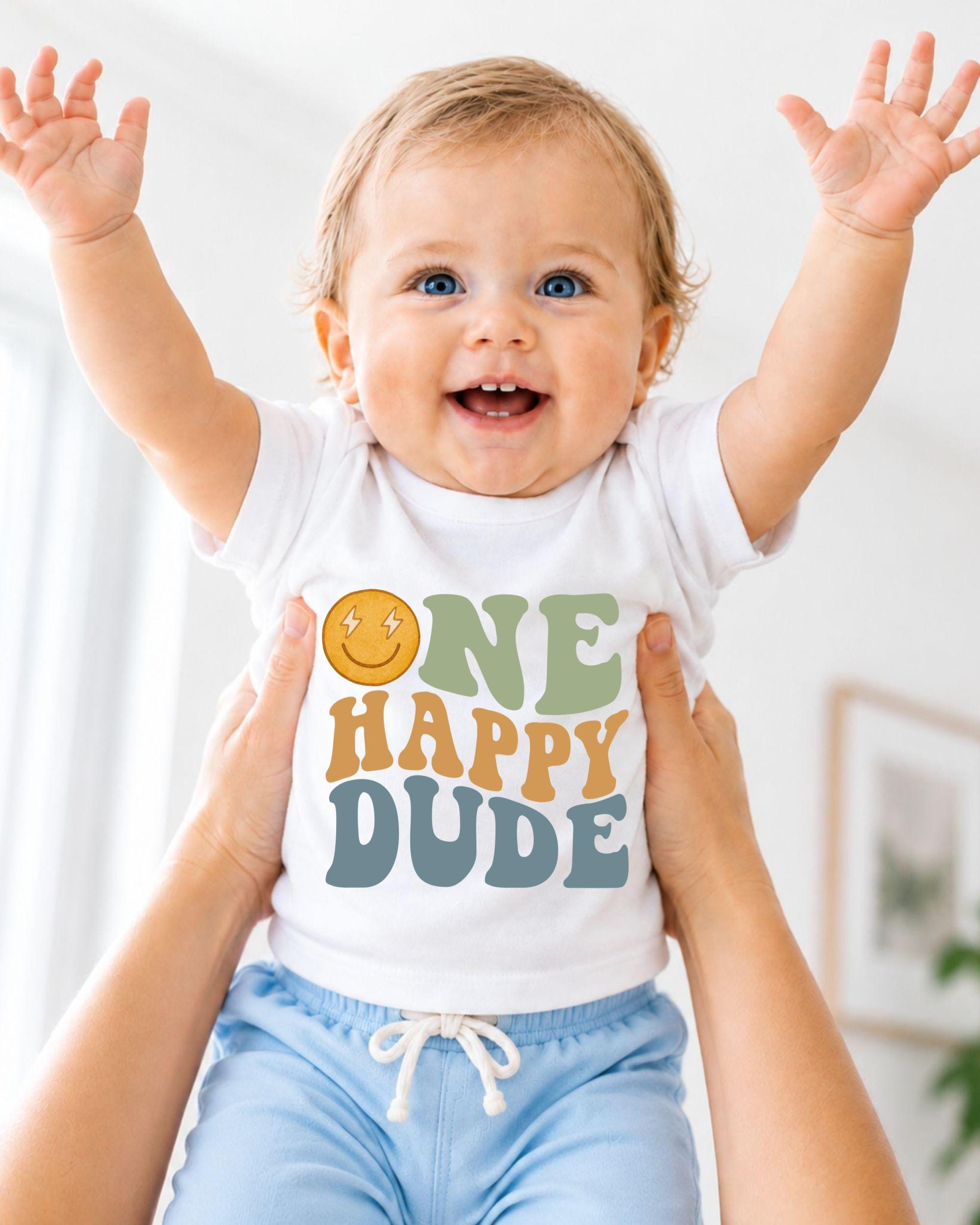 A smiling baby wearing a white shirt with the text "ONE HAPPY DUDE" is being held up by someone's arms.
