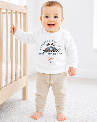 A smiling baby boy wearing a white sweatshirt with a racing theme and text, standing in a nursery setting next to a crib.