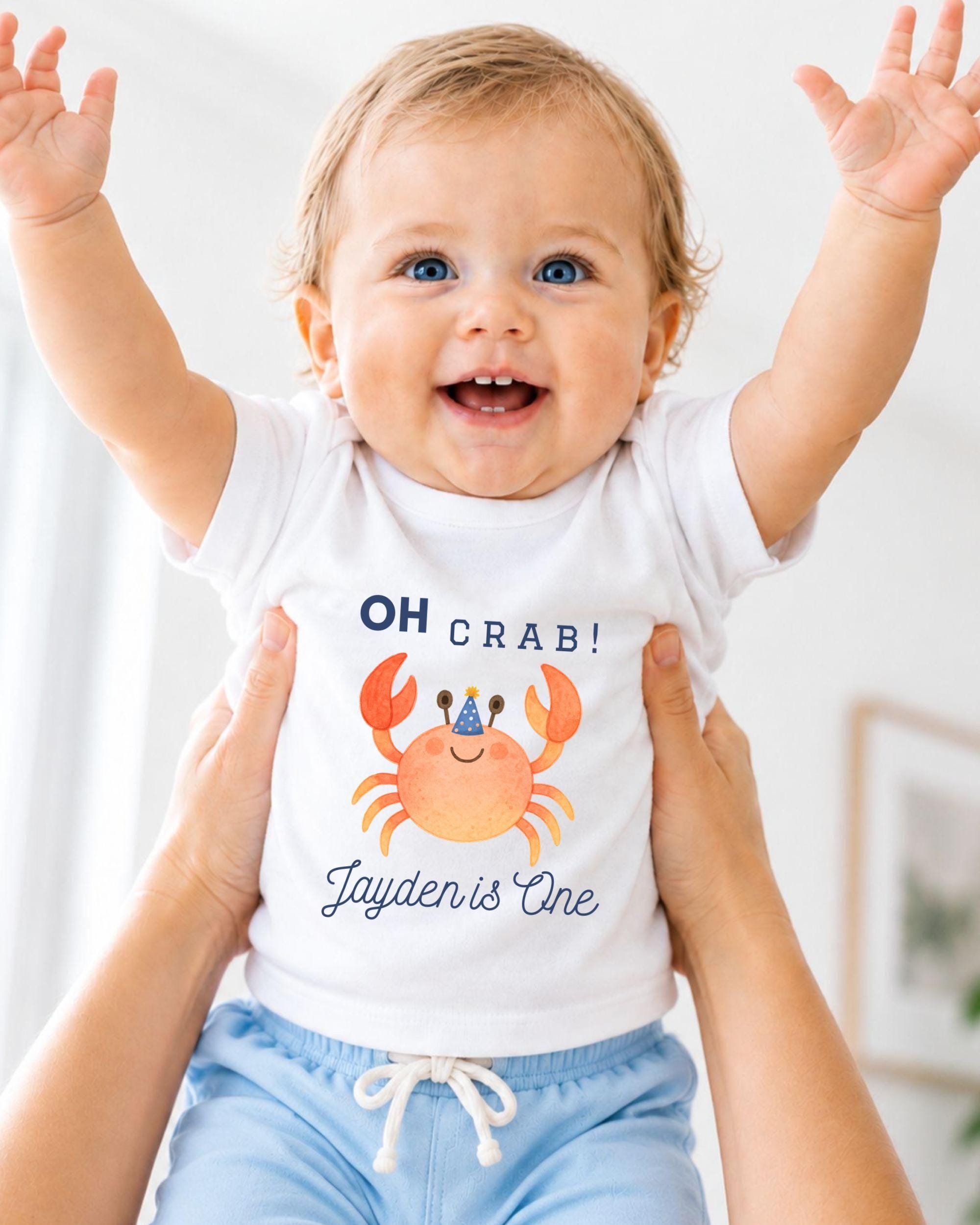 A smiling baby wearing a white t-shirt with a crab design and the text "OH Crab! Jayden is One" is being held up by someone's arms.