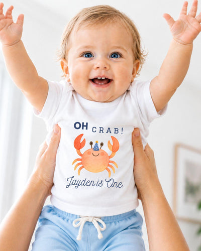 A smiling baby wearing a white t-shirt with a crab design and the text "OH Crab! Jayden is One" is being held up by someone's arms.
