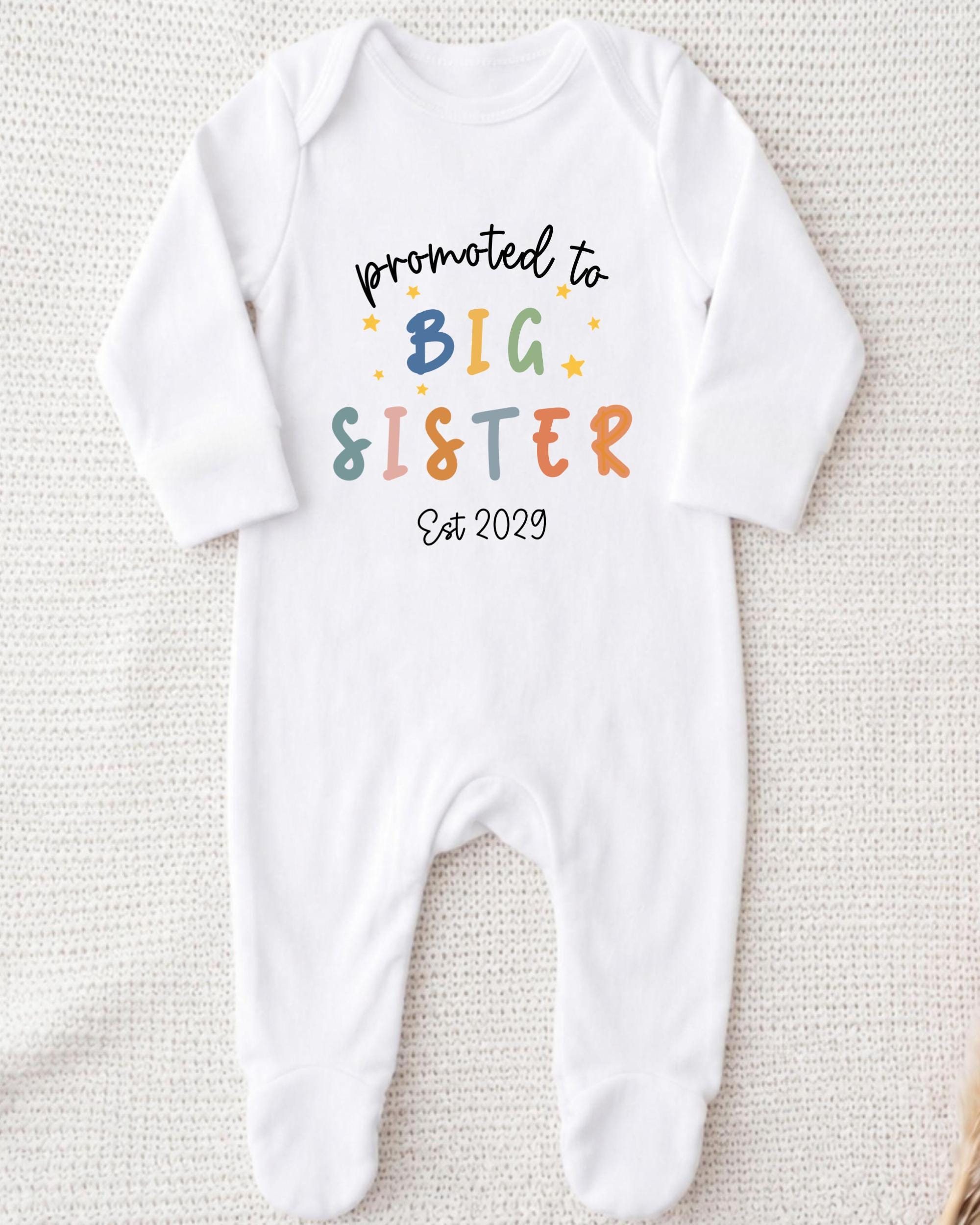 A white onesie with text that says "promoted to BIG SISTER Est 2029" printed on it.