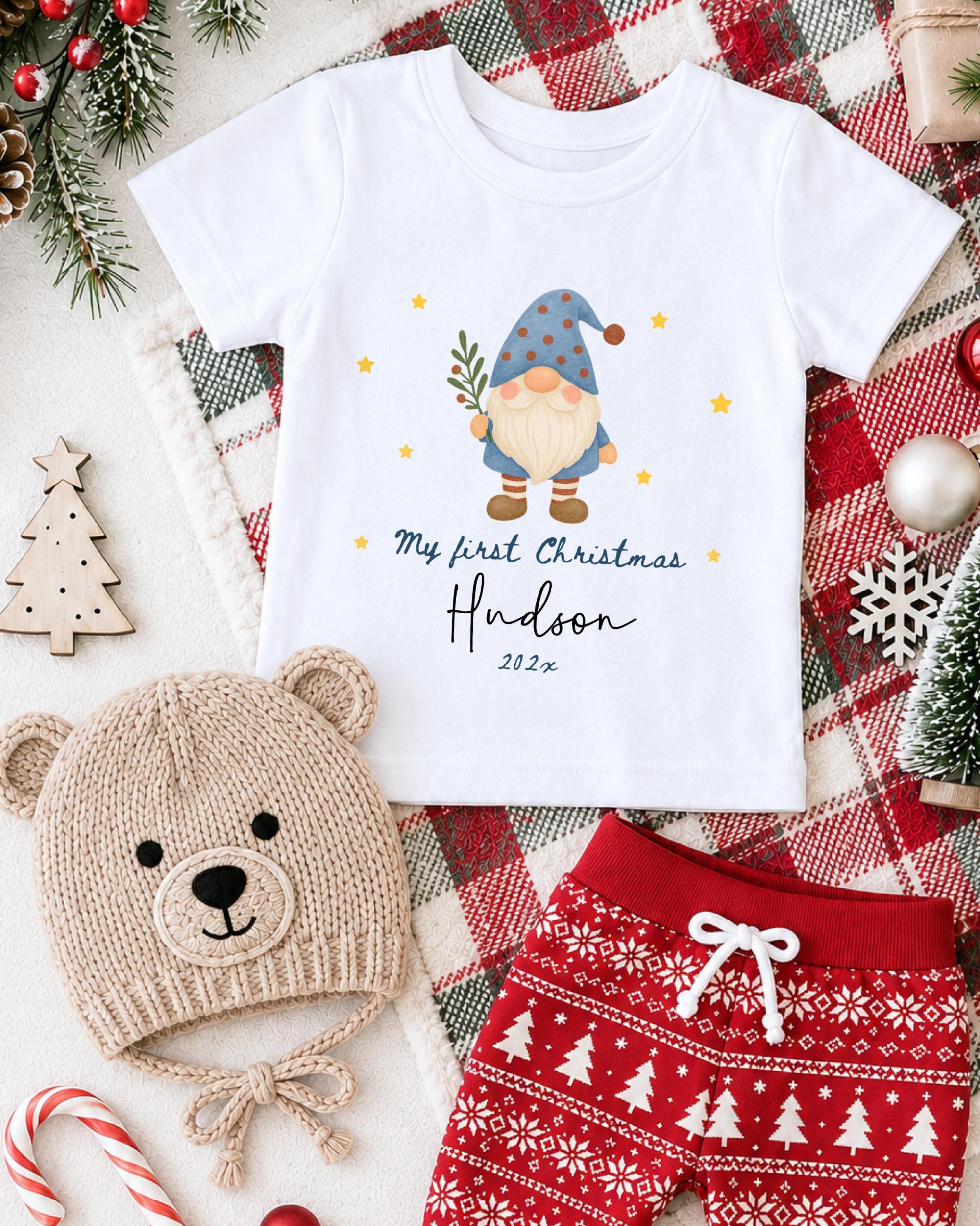 A white t-shirt with a gnome graphic and the text "My first Christmas Hudson" printed on it, along with a red and white patterned pair of shorts, a beige knit hat with a gnome face, and Christmas decorations such as a tree, ornaments, and a snowflake.