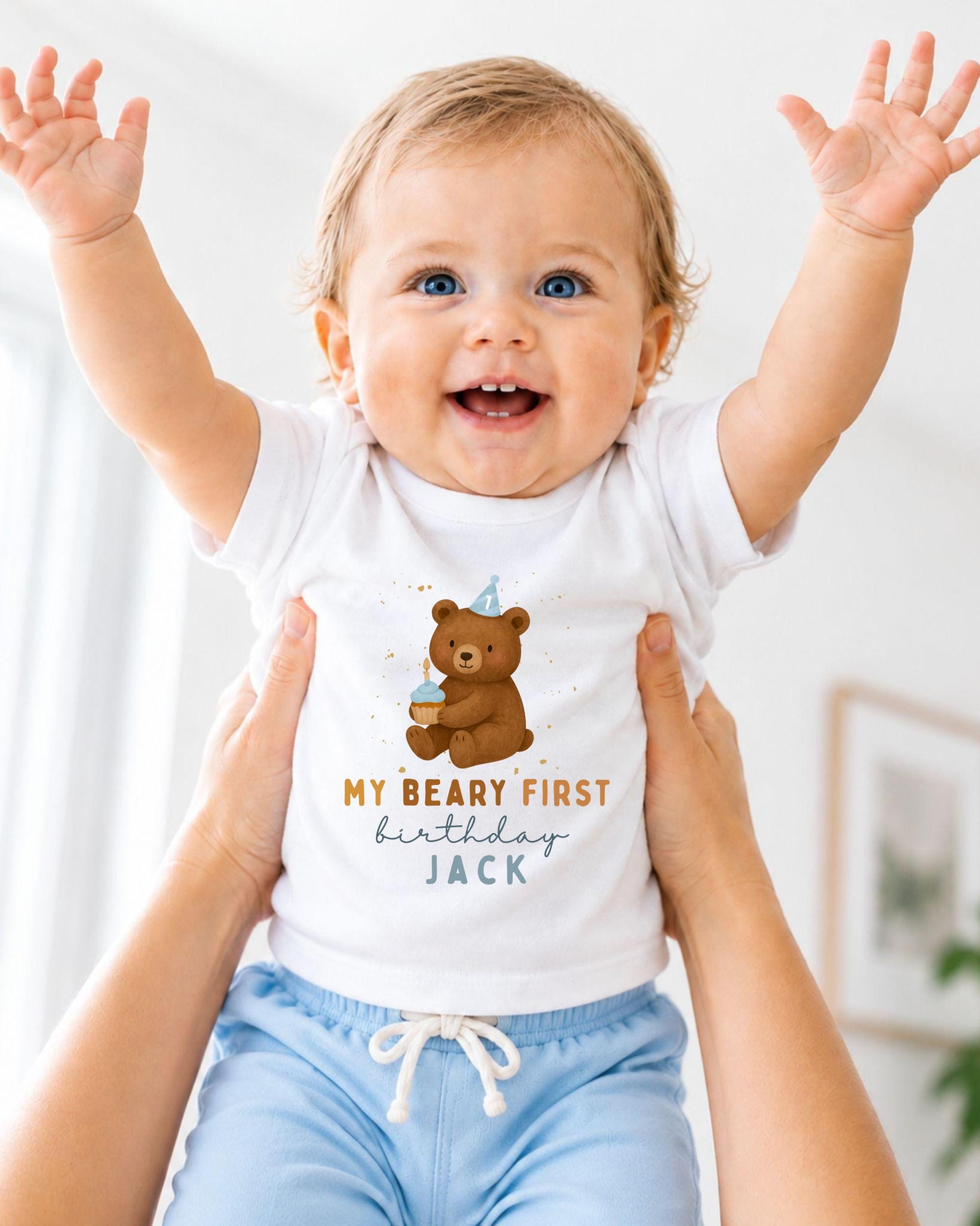 A smiling baby wearing a white t-shirt with a teddy bear and the text "My Beary First birthday Jack" is being held up by someone's arms.