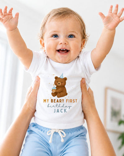 A smiling baby wearing a white t-shirt with a teddy bear and the text "My Beary First birthday Jack" is being held up by someone's arms.