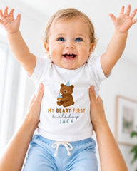 A smiling baby wearing a white t-shirt with a teddy bear and the text "My Beary First birthday Jack" is being held up by someone's arms.