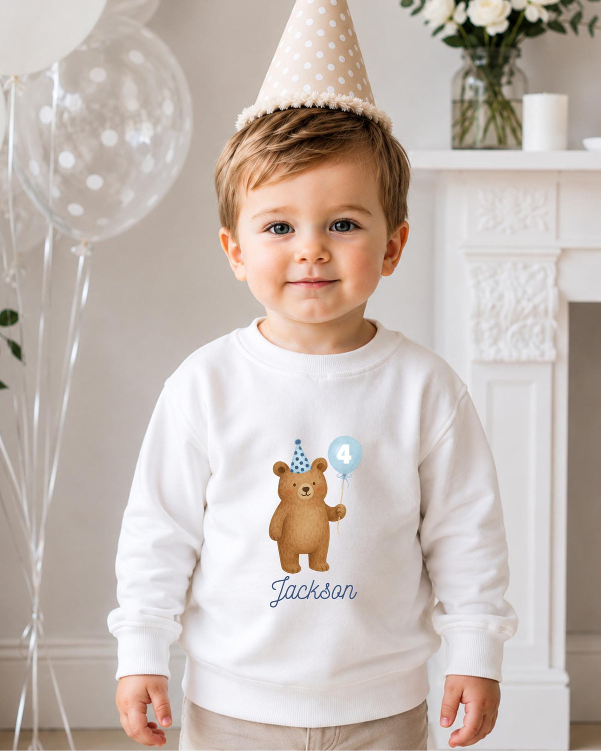 A young boy wearing a white sweatshirt with a teddy bear and a birthday hat, smiling at the camera.
