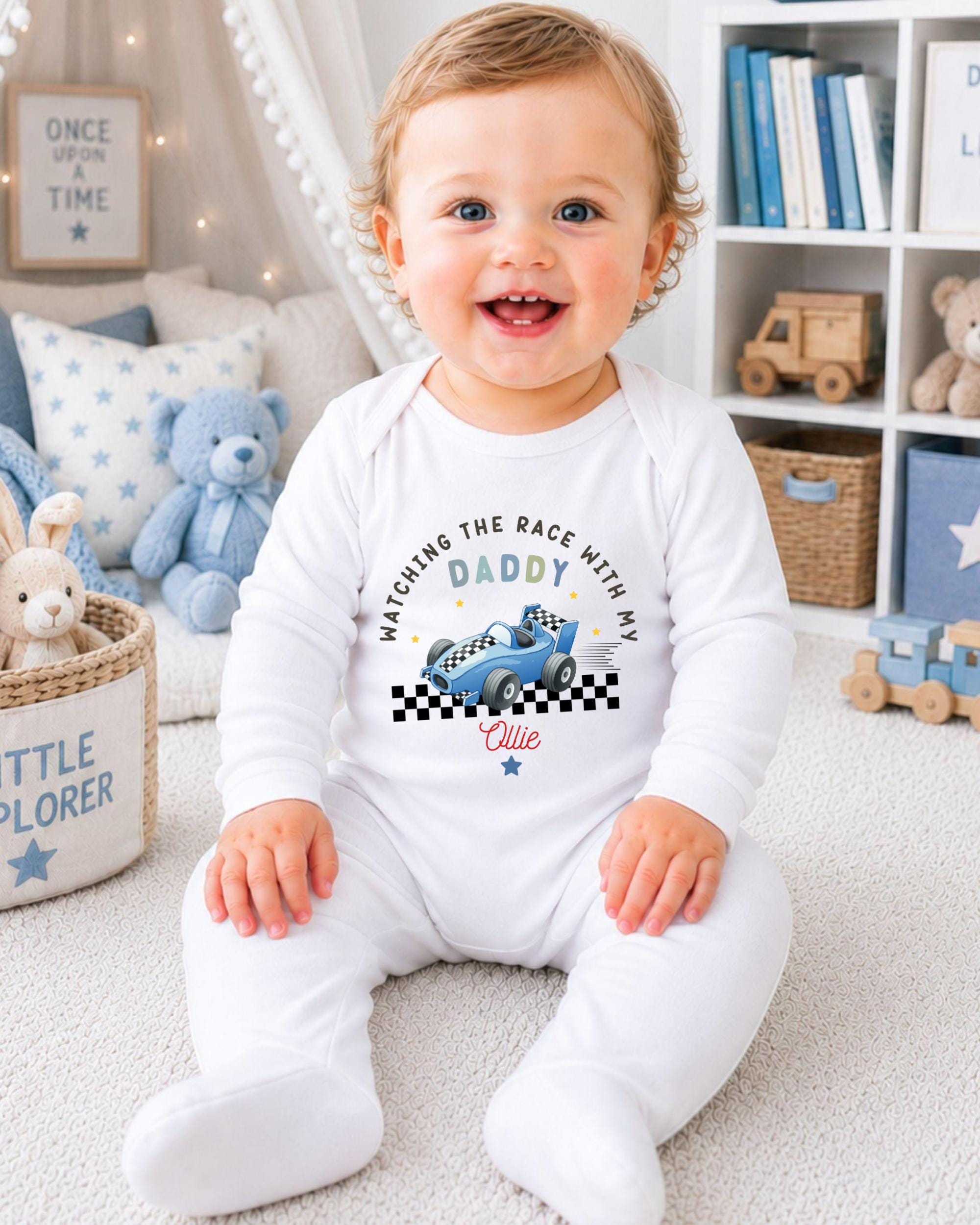 A smiling baby boy wearing a white onesie with a racing car design sits on the floor in a nursery setting.