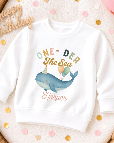 A white sweatshirt with a colorful graphic of a whale and balloons, along with the text "One-DER The Sea Harper" printed on it.