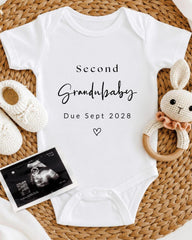 A white onesie with the text "Second Grandbaby Due Sept 2028" printed on it, along with a crocheted rabbit toy and a black and white photograph of an infant's belly.