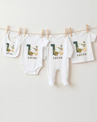A collection of white baby clothes, including onesies and pajamas, with the number 1 and dinosaur designs printed on them.