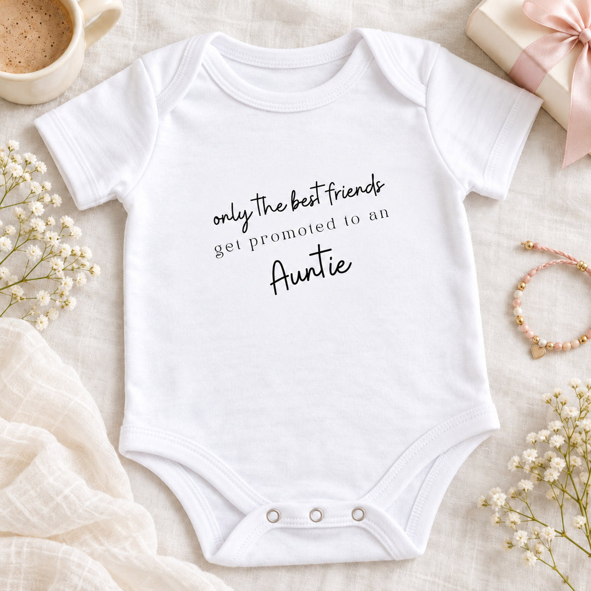 Square pregnancy announcement image of a white Best Friends Promoted to Auntie Baby Vest, styled on beige linen with a coffee cup, gift box, ribbon and friendship bracelet.