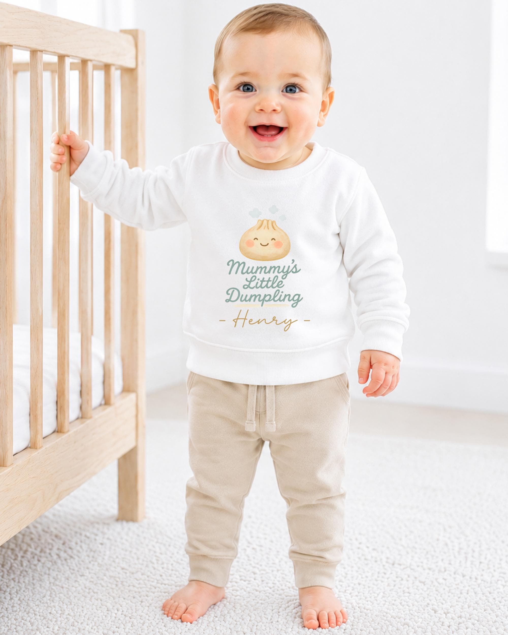 A smiling baby boy wearing a white sweatshirt with a cartoon character and text printed on it, standing next to a wooden crib.