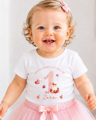 A smiling baby girl wearing a white t-shirt with a number 1 and pink flowers, a pink tutu skirt, and a pink bow in her hair.