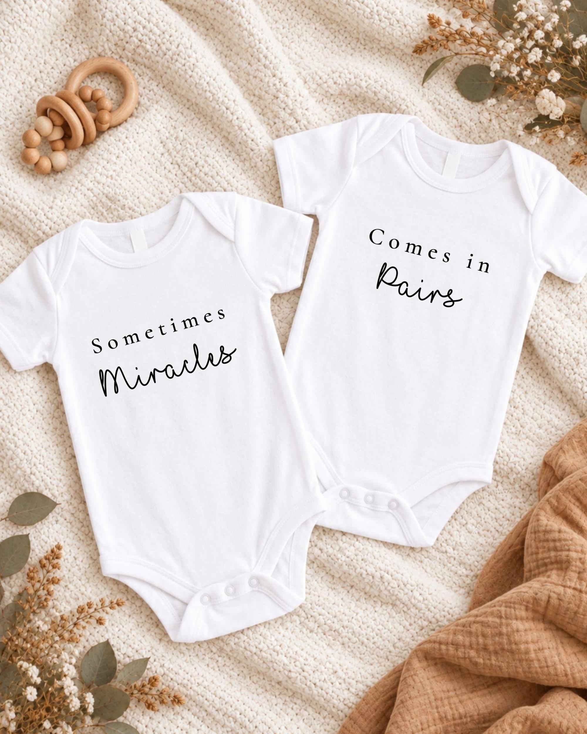 two white baby onesies with the text "Sometimes Miracles" printed on them, placed on a white blanket with some floral decorations.