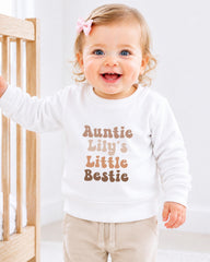 A smiling baby girl wearing a white sweater with the text "Auntie Lily's Little Bestie" printed on it.