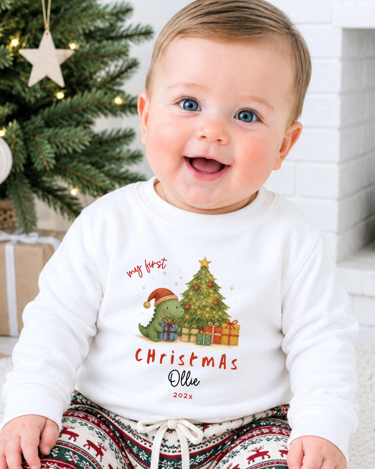 A smiling baby wearing a white Christmas-themed sweatshirt with a dinosaur and presents, sitting in front of a Christmas tree.