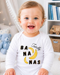 A smiling baby wearing a white onesie with a banana graphic and the text "Totally Bananas" is sitting in a room with a teddy bear and bookshelf in the background.