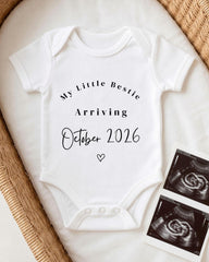 A white onesie with text announcing the arrival of a baby in October 2026, placed on a bed with a woven basket.