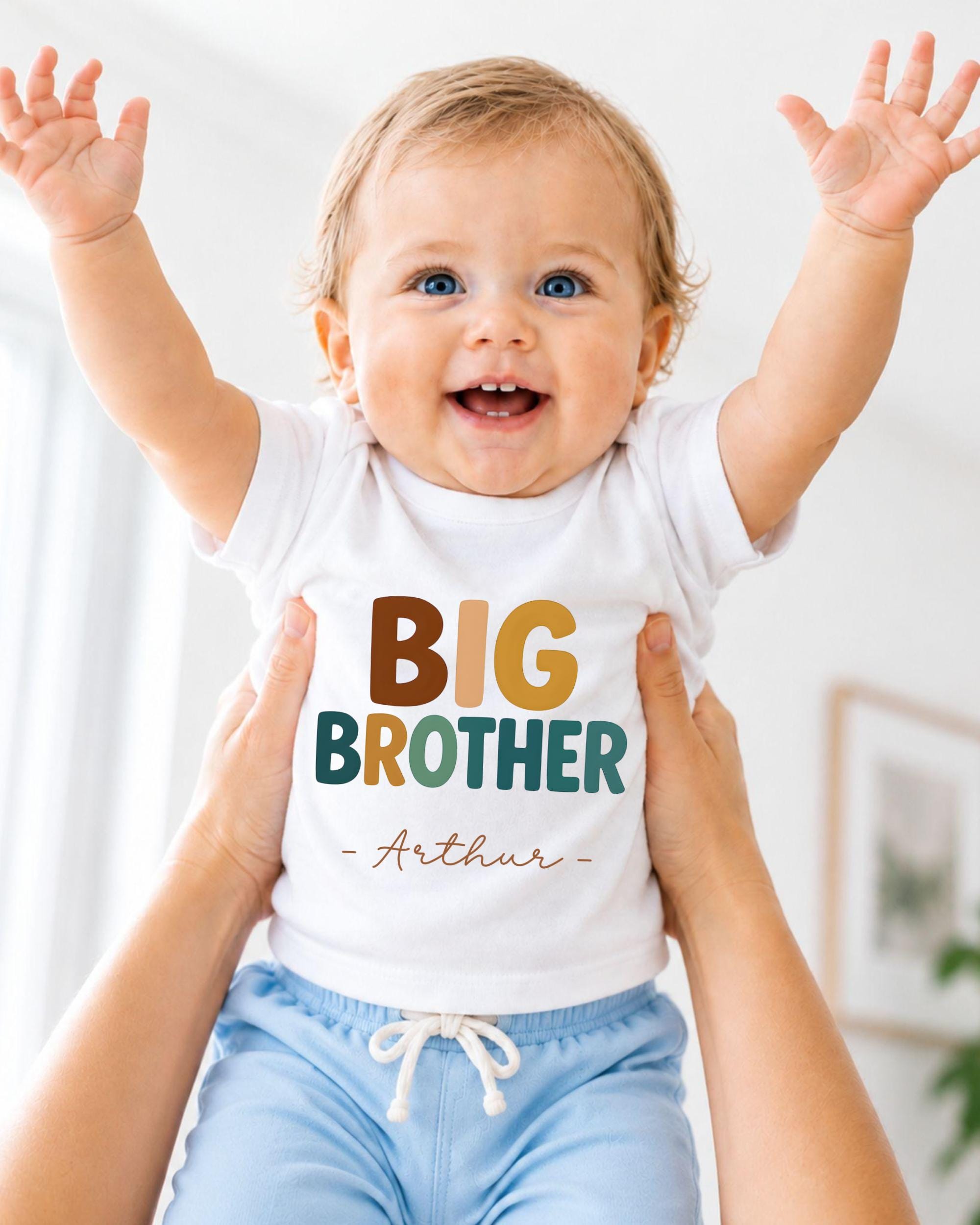 A smiling baby wearing a white shirt with the text "BIG BROTHER - ARTHUR" is being held up by someone's arms.