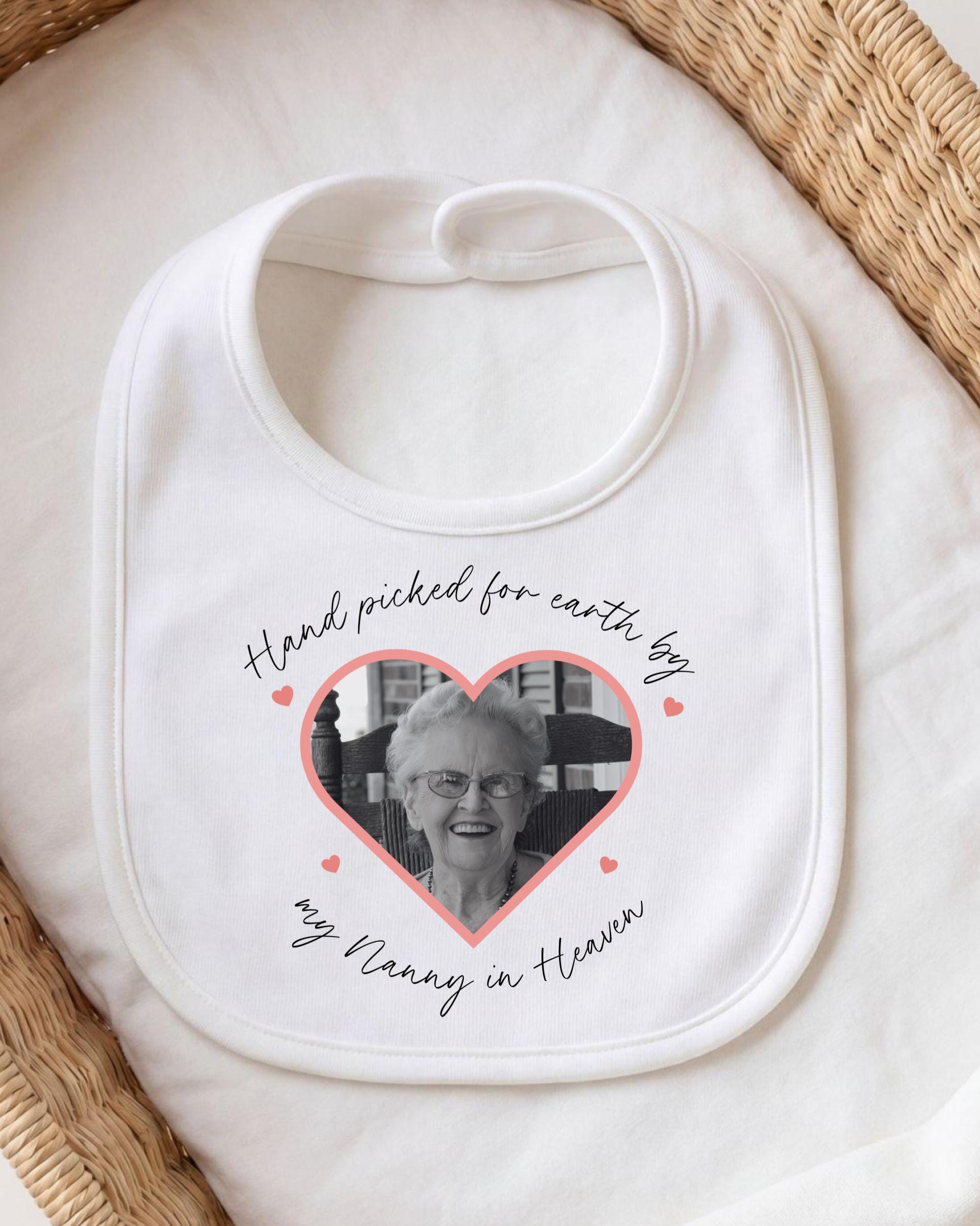 A white bib with a photo of an elderly woman inside a heart shape, surrounded by pink hearts. The text on the bib reads "Hand picked for earth by my Nanny in Heaven".